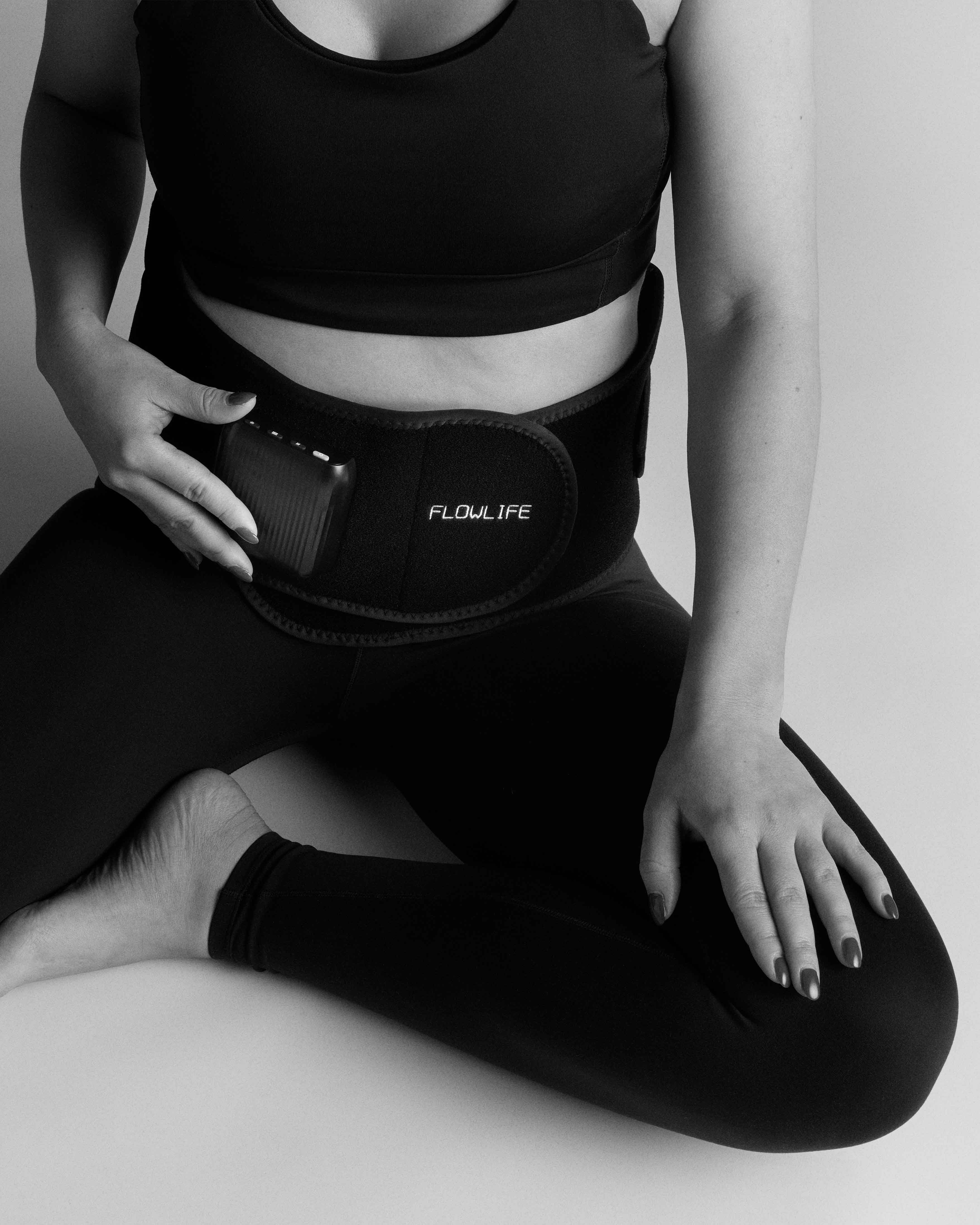 A woman wearing the black Flowtherma Belt for lumbar heat and vibration therapy while sitting on the floor.